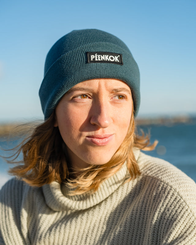 PÉENKOK’ | ORGANIC COTTON BEANIE TEAL WITH A WOVEN PATCH IN RECYCLED POLYESTER EMBROIDERY ON THE FRONT WORN BY A WOMAN AT THE BEACH